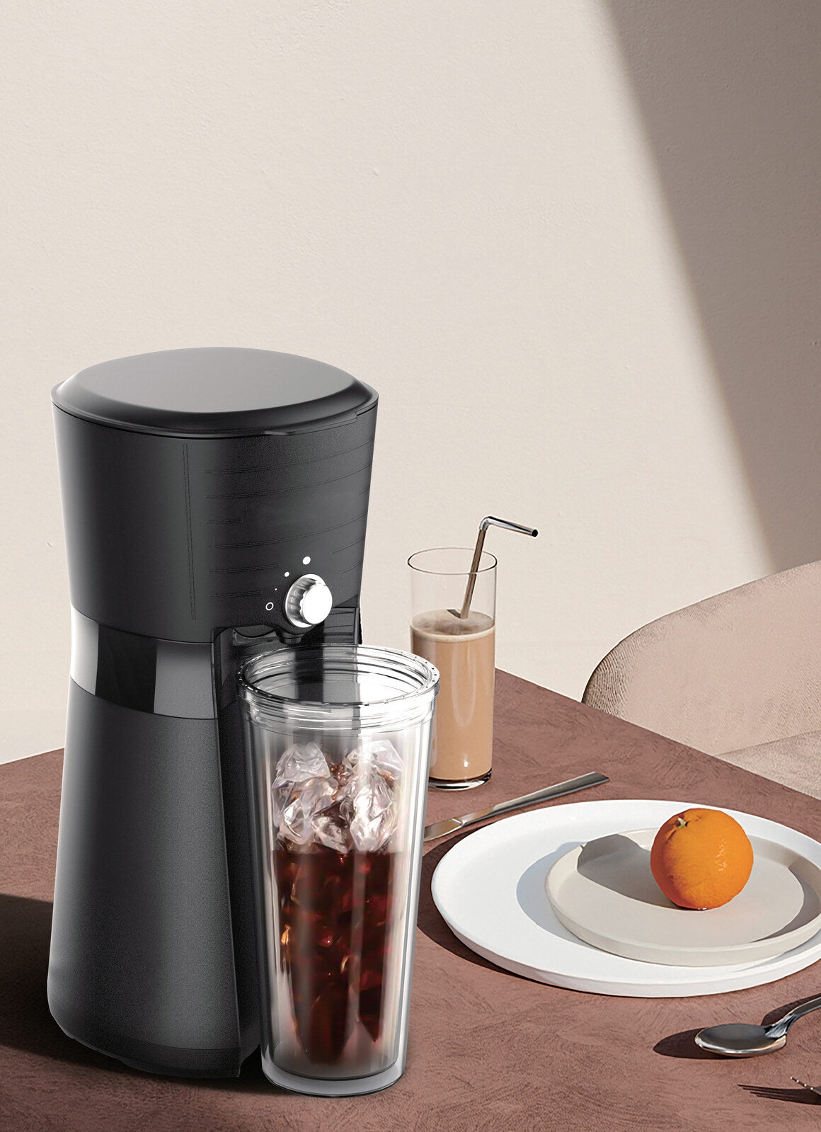 Black coffee maker on a table with a glass of iced coffee and a plate with an orange.