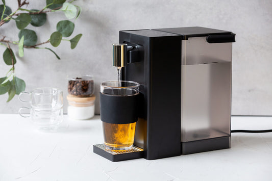 Modern coffee machine dispensing a glass of coffee on a light surface with a plant in the background.