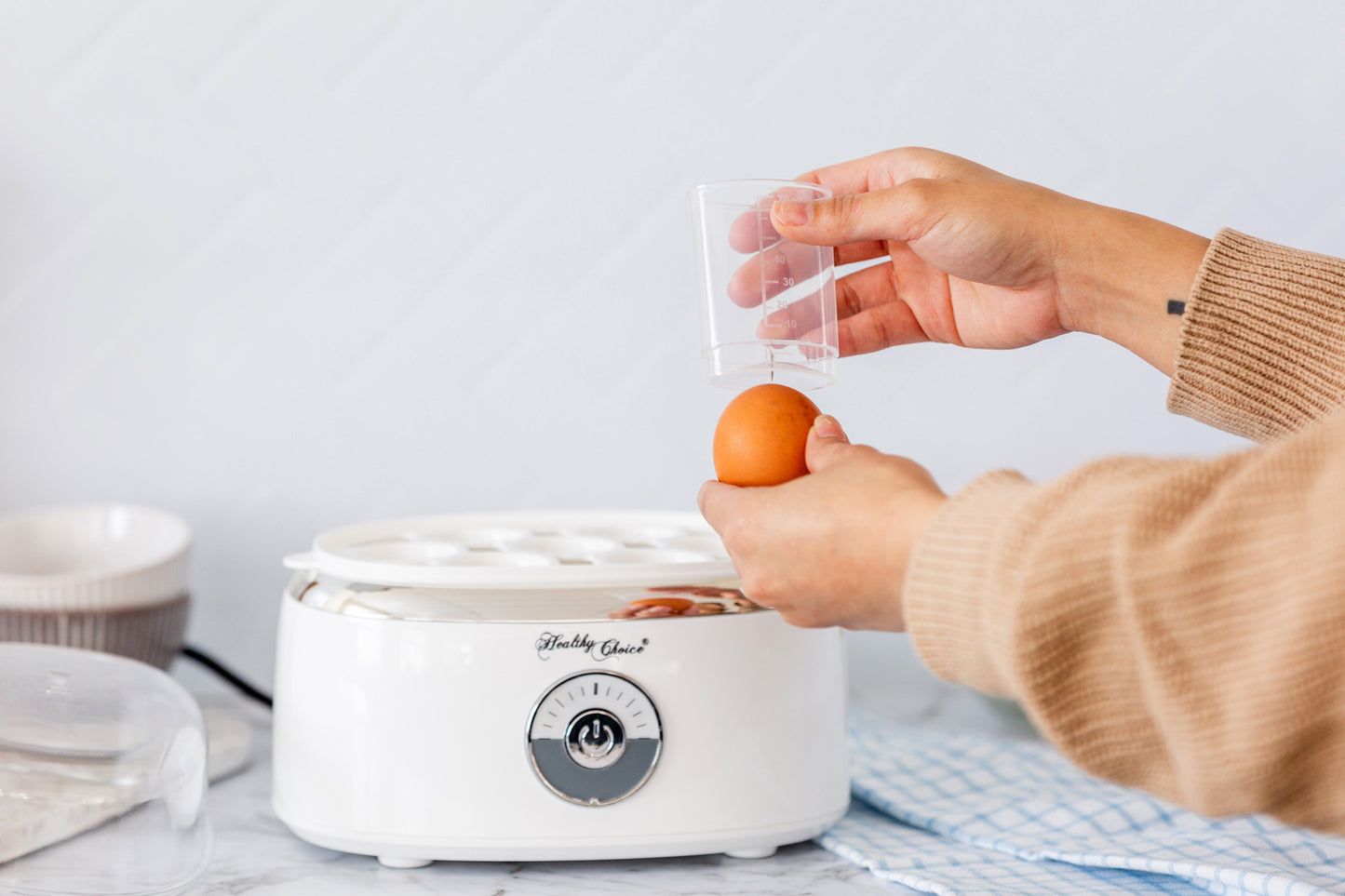 Person using a small white appliance with a brand logo, likely an egg cooker, on a light-colored surface.