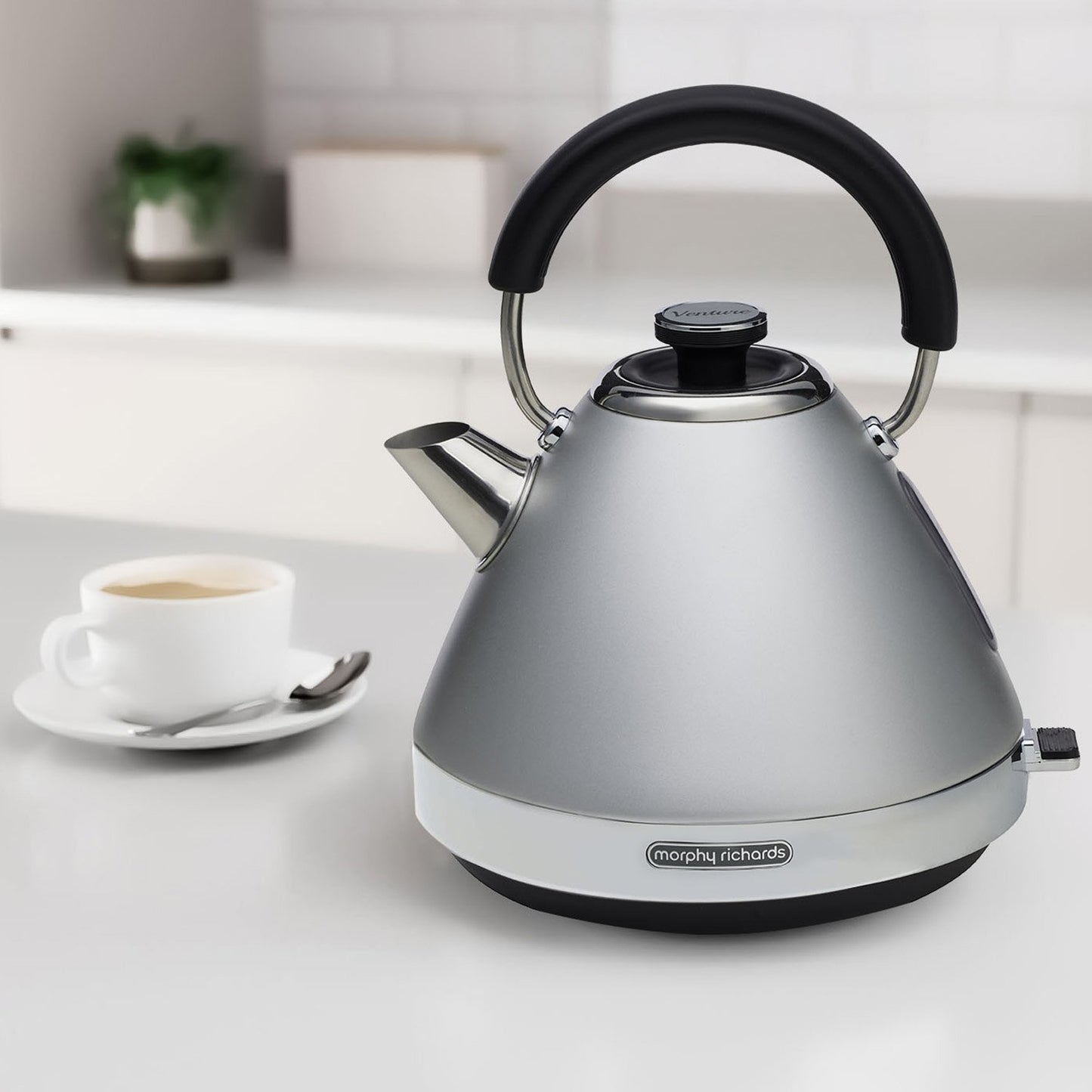Silver kettle with black handle on a kitchen counter with a cup of coffee in the background