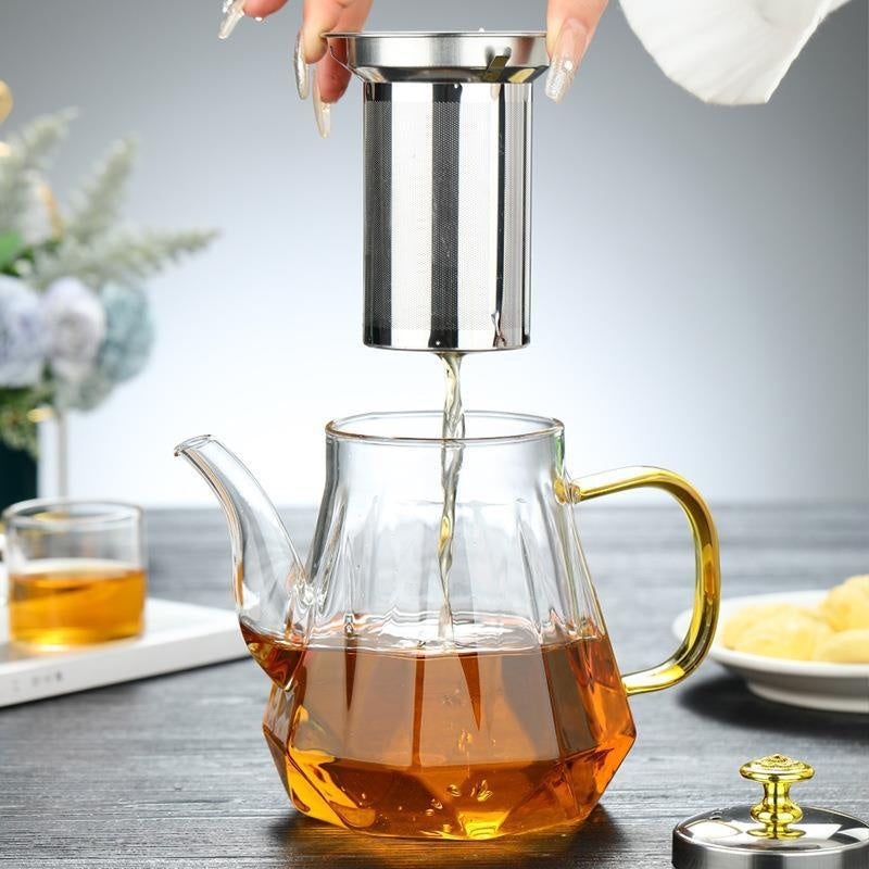 Glass teapot with metal infuser being filled with tea on a wooden table.