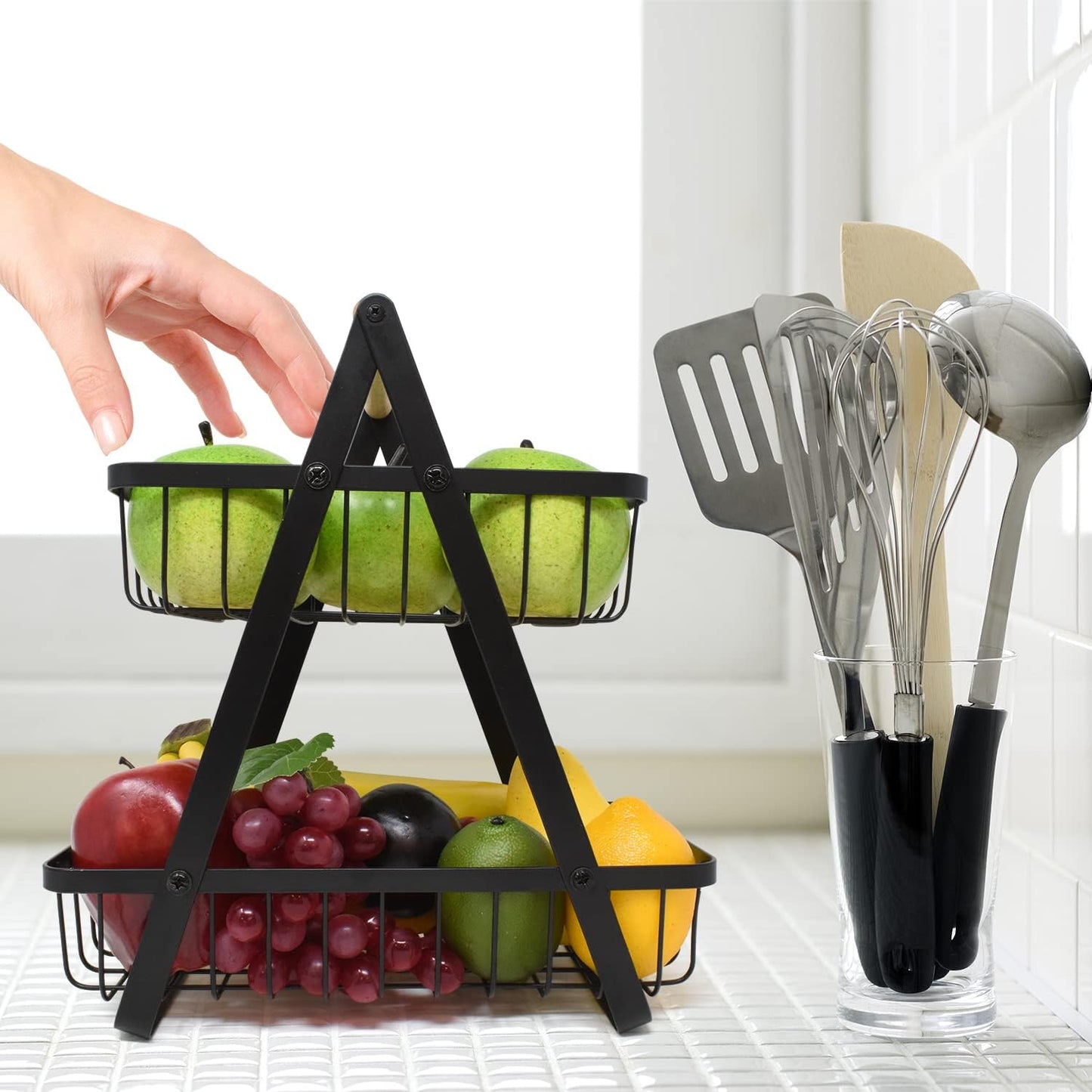 Two-tier fruit basket with apples and grapes on a kitchen counter, next to kitchen utensils.