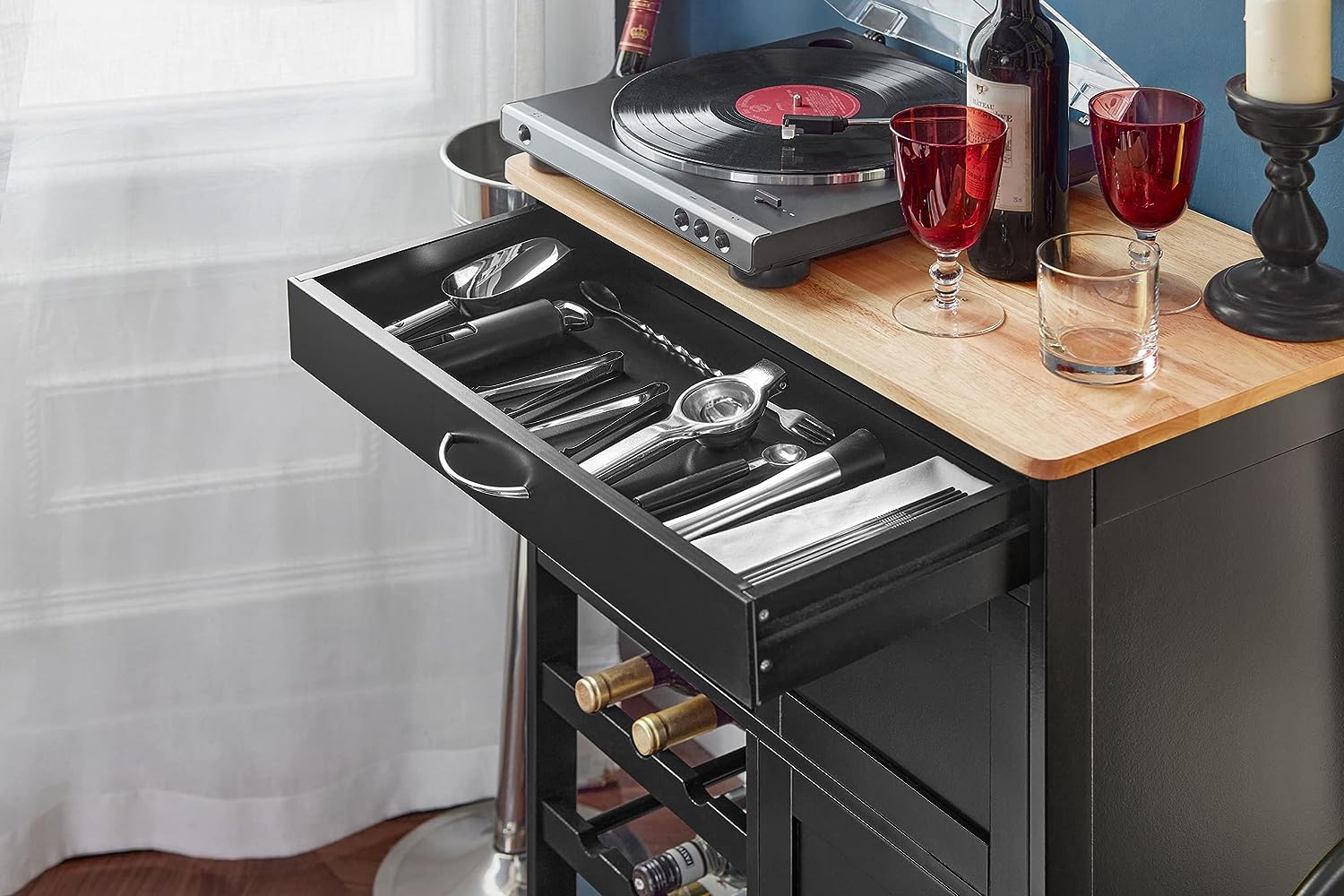 Black kitchen island with open drawer displaying silverware, wine bottles, and glasses on a wooden surface.