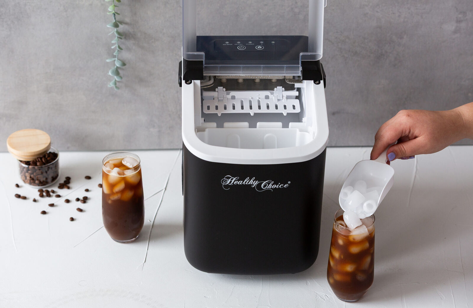 Ice cube maker with a hand adding ice to a glass of iced coffee on a white surface.