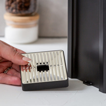 Person holding a small metal device with a grid pattern next to a black appliance.