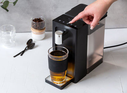 Black coffee machine dispensing a glass of coffee on a white countertop.