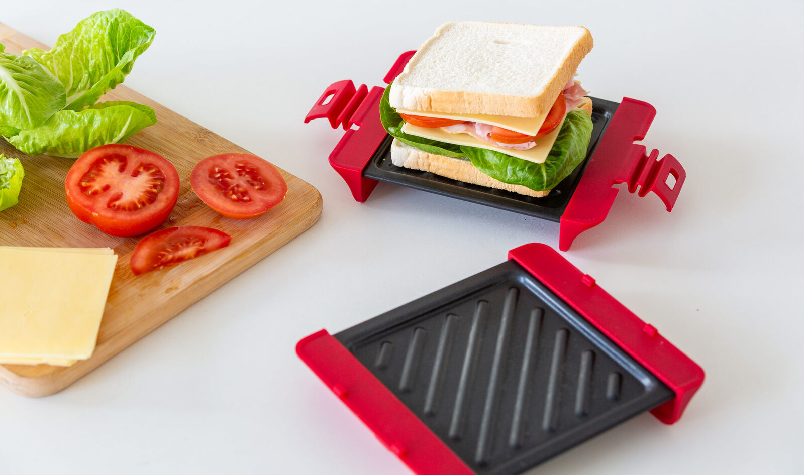 Red sandwich maker with a sandwich being made, surrounded by ingredients on a white surface.