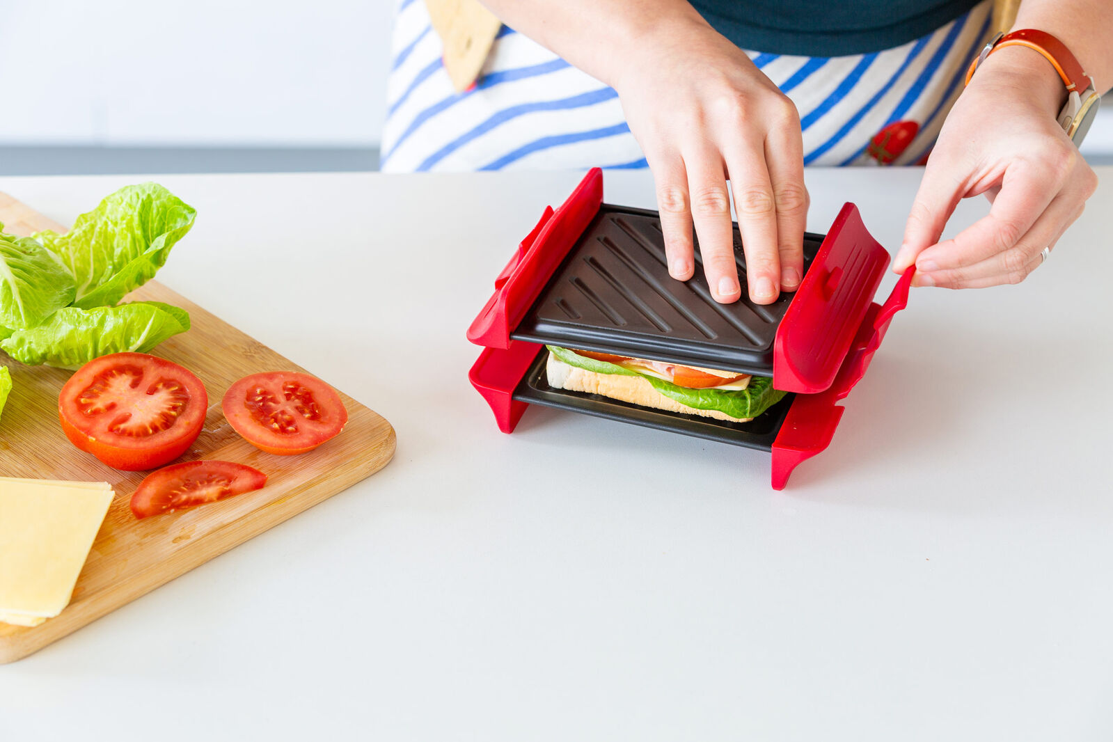 Person making a sandwich using a red and black sandwich maker on a white surface with ingredients nearby.