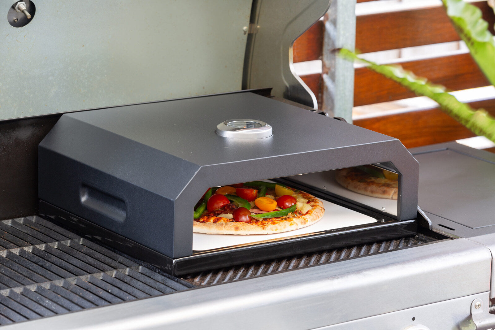 Portable pizza oven on a grill with a pizza inside, surrounded by outdoor elements.