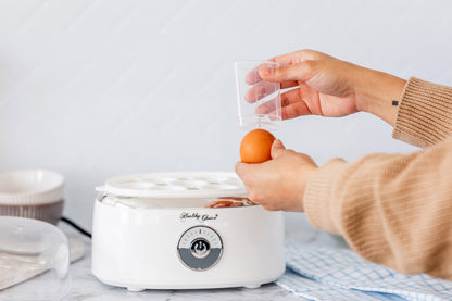 Person using a small white appliance with a brand logo, likely an egg cooker, on a light-colored surface.