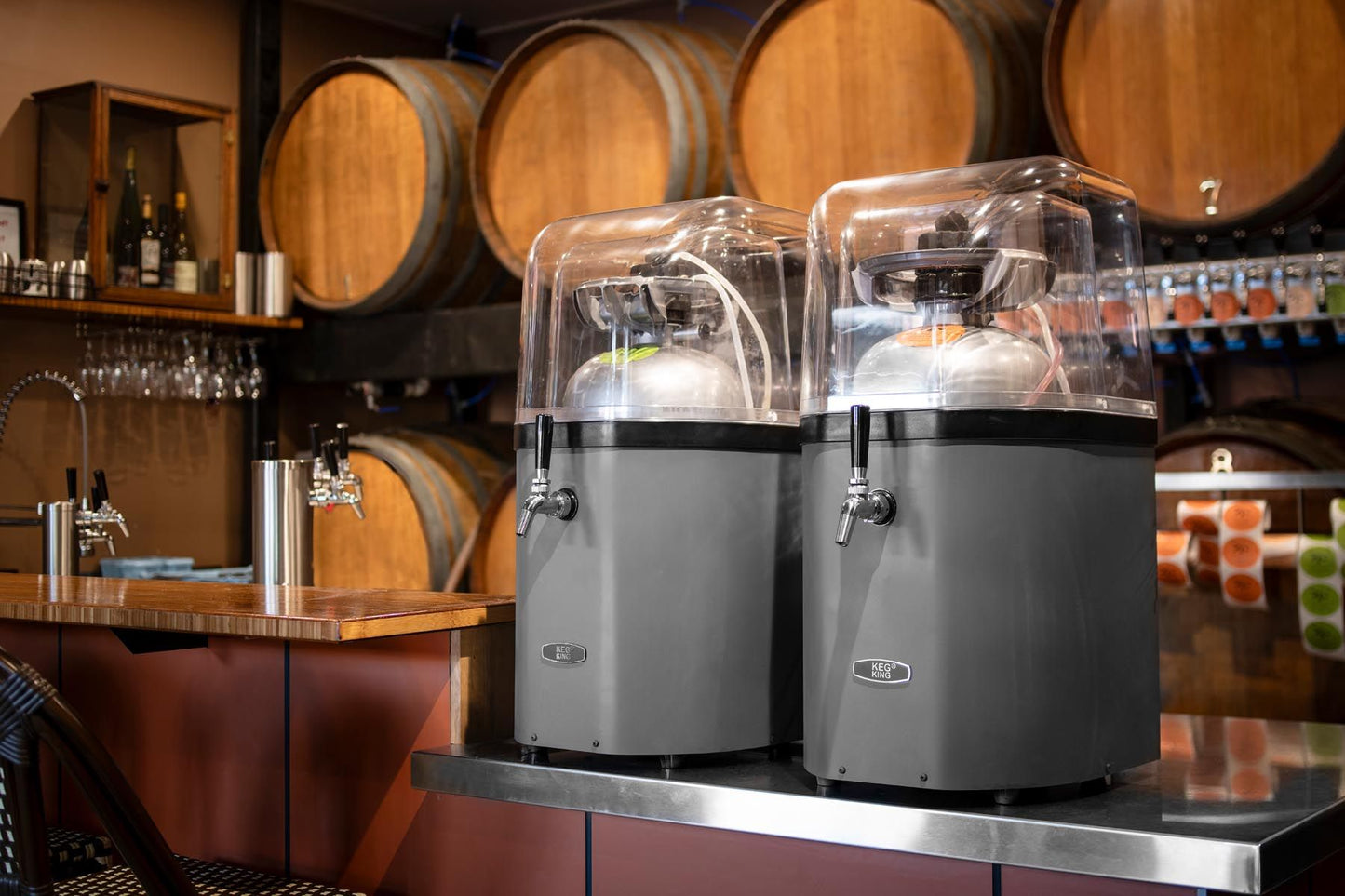 Two large black dispensers on a bar counter with wooden barrels in the background.