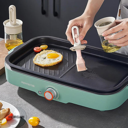 Person using a brush to apply oil on a grill with food, next to a bottle of oil and a cup.