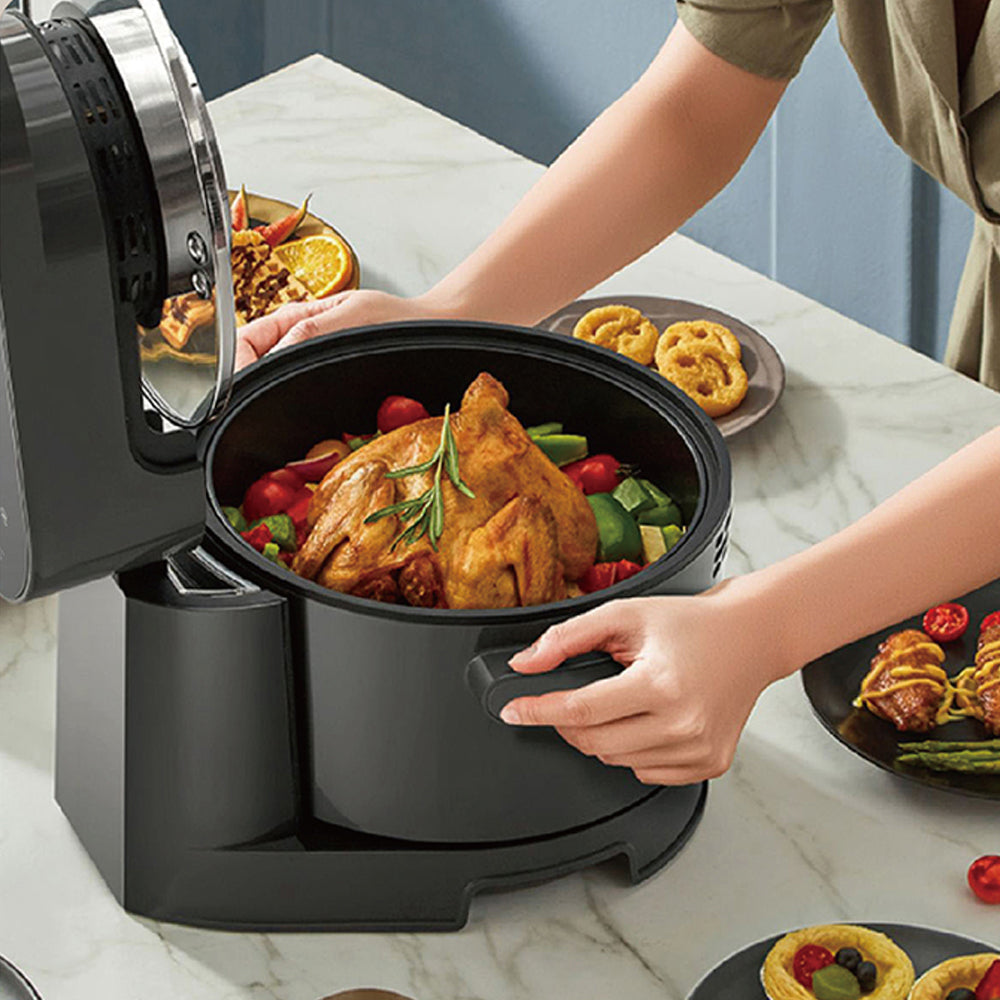 Person opening an air fryer to reveal a roasted chicken with vegetables on a marble countertop.